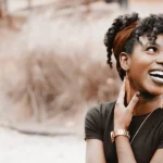 WELL SAID: TORONTO SPEECH THERAPY. A woman with curly hair and a headband smiles brightly, looking to the side. She wears a black top, gold hoop earrings, and a gold watch, with one hand touching her neck. The softly blurred background hints at an Ontario setting.
