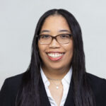 WS-0313print WELL SAID: TORONTO SPEECH THERAPY. A person with long dark hair and glasses is smiling, wearing a white shirt, a black blazer, and layered necklaces, against a plain light background.