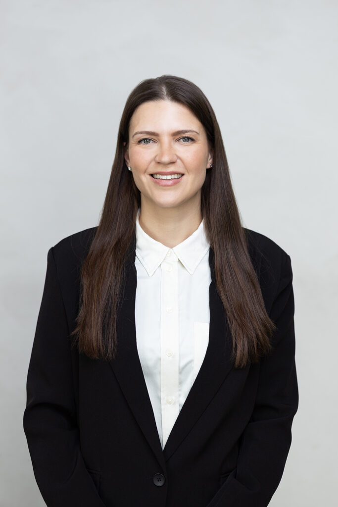 WELL SAID: TORONTO SPEECH THERAPY. A woman with long straight brown hair wearing a black blazer over a white button-up shirt stands in front of a plain light background in Toronto, smiling at the camera.