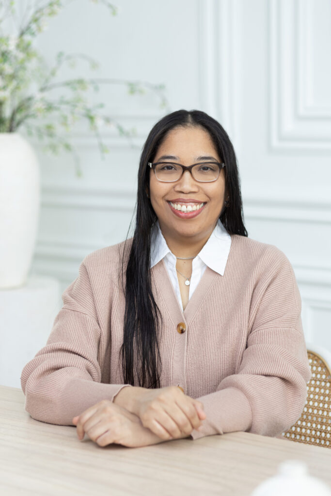 WELL SAID: TORONTO SPEECH THERAPY. A woman with long dark hair and glasses sits at a table in Toronto, smiling at the camera. She is wearing a light pink cardigan over a white collared shirt. A vase with green branches is in the background.
